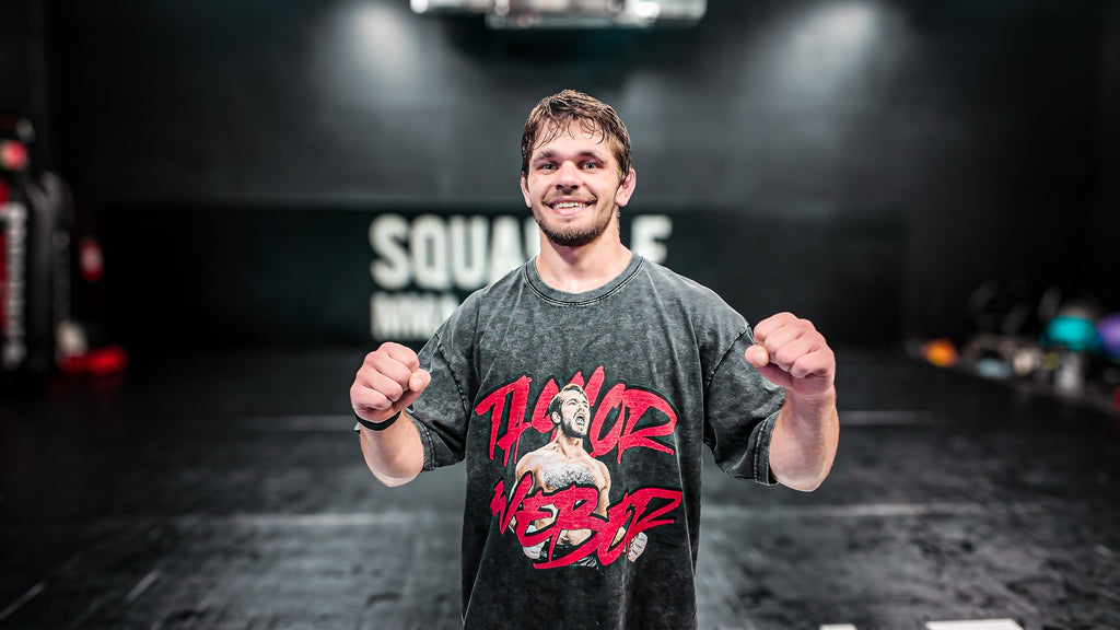 Person wearing a gray t-shirt with red graphics and text, standing in a dark indoor setting.